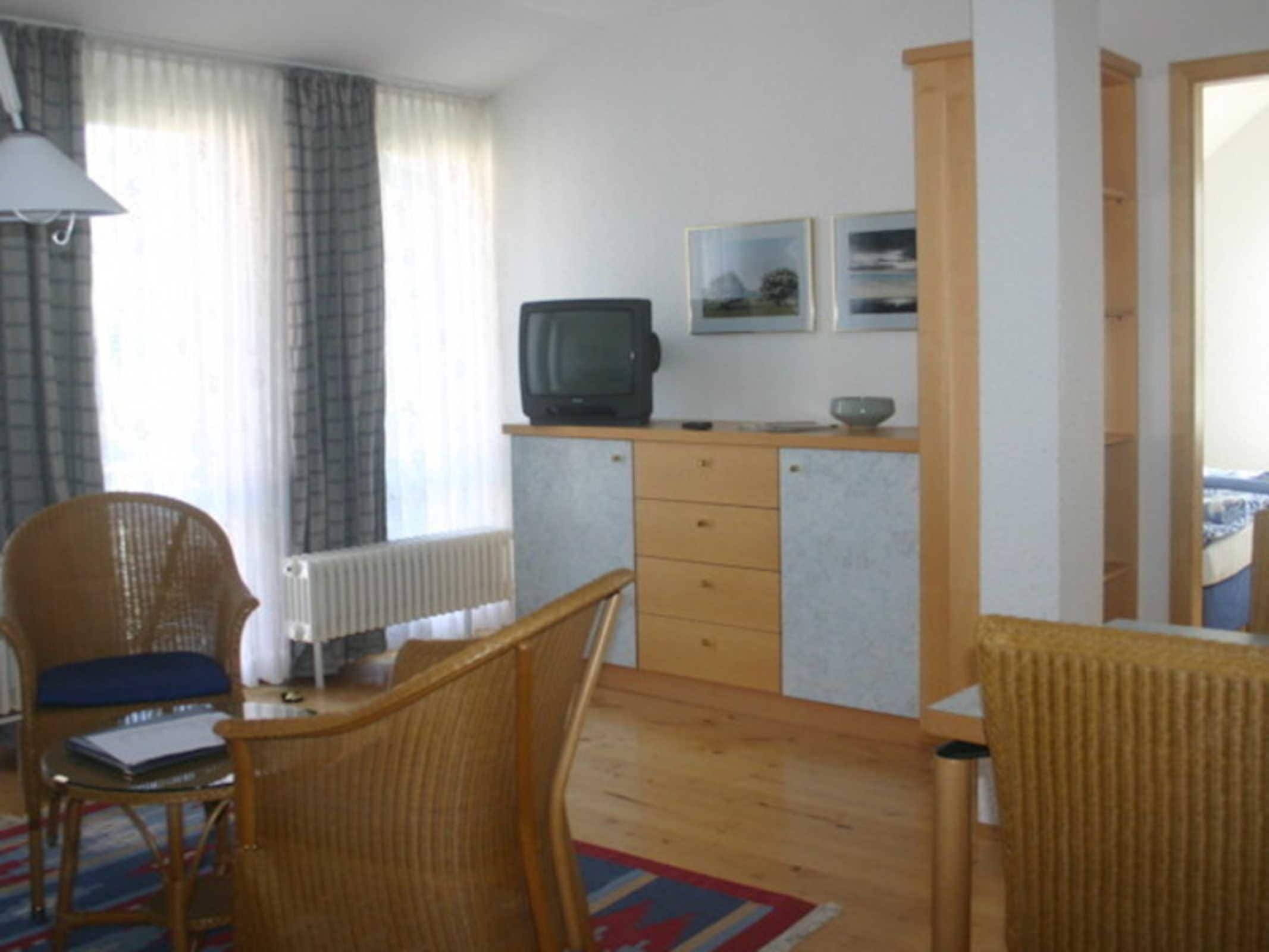 A room with a wooden desk, two wicker chairs, a radiator, a television, and two framed pictures on the wall.