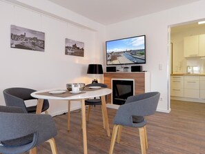 A modern living room with a dining table, grey chairs, a flat-screen TV mounted above a fireplace, and a kitchen visible through an open doorway. - Ohlerich storage app. 36 - Ohlerich Speicher App. 36 (Wismar)