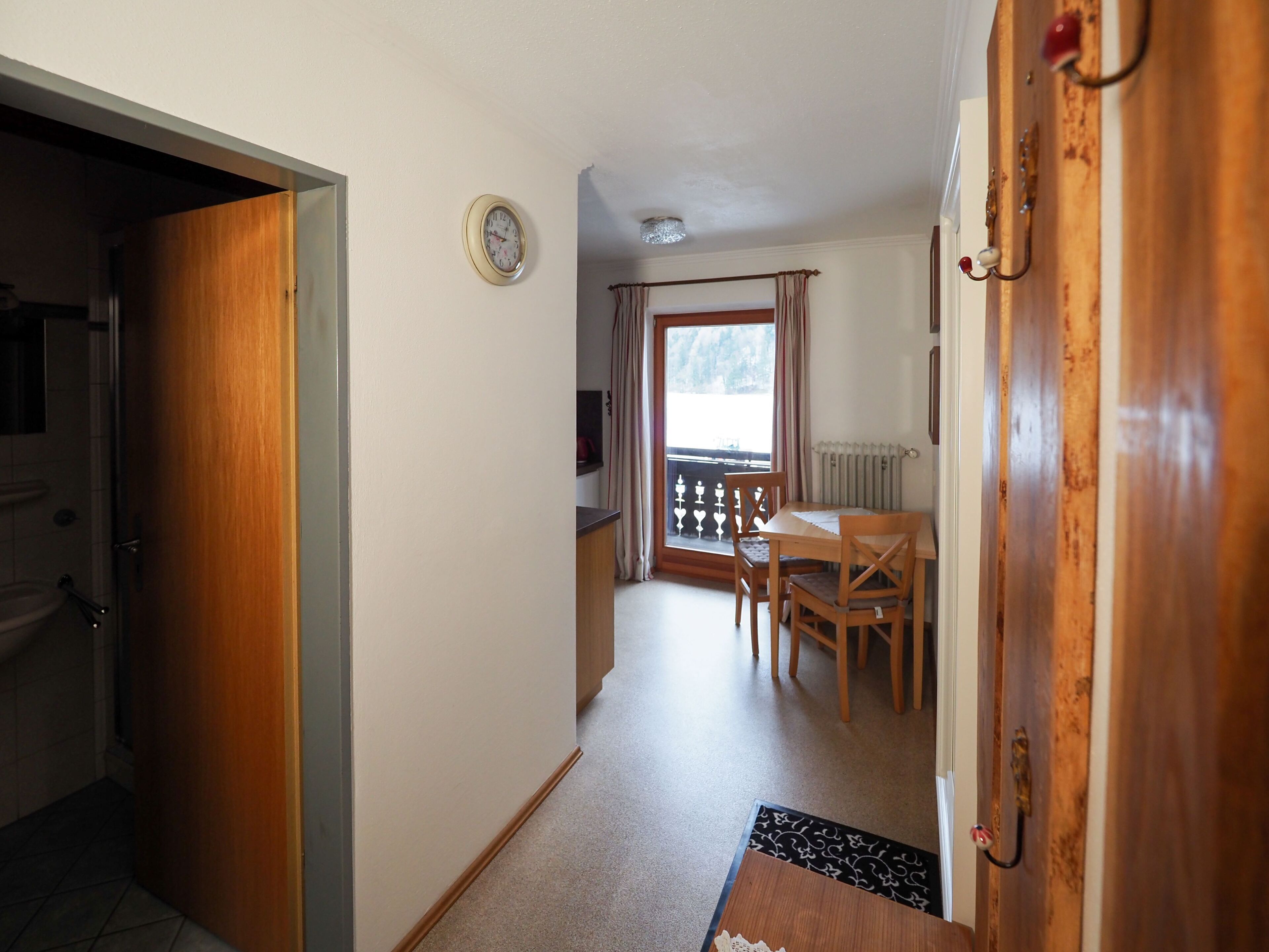 A hallway leading to a dining area with a wooden table and chairs, a clock on the wall, and a view through a window with curtains.