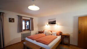 A bedroom with a wooden bed, a bench, a window with curtains, a radiator, a wall-mounted picture, and a door. - Apartment Geigelstein for 2 pers., 36 sqm, balcony (Schleching)