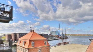 Blick auf das Wasser mit einem Backsteinhaus, einem Steg mit Booten und Industriekranen.