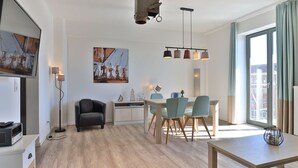 En moderne stue med spiseplads, en sort lædersofa, et træspisebord og et stort vindue med gardiner.