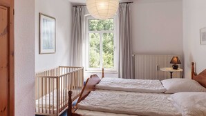 Et soveværelse med en træseng, en barneseng, et vindue med gardiner, en radiator og en lampe på et sengebord.