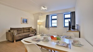 Un salon moderne avec un canapé, une table à manger et des chaises. Il y a une fenêtre avec des rideaux, une lampe et un tableau au mur.