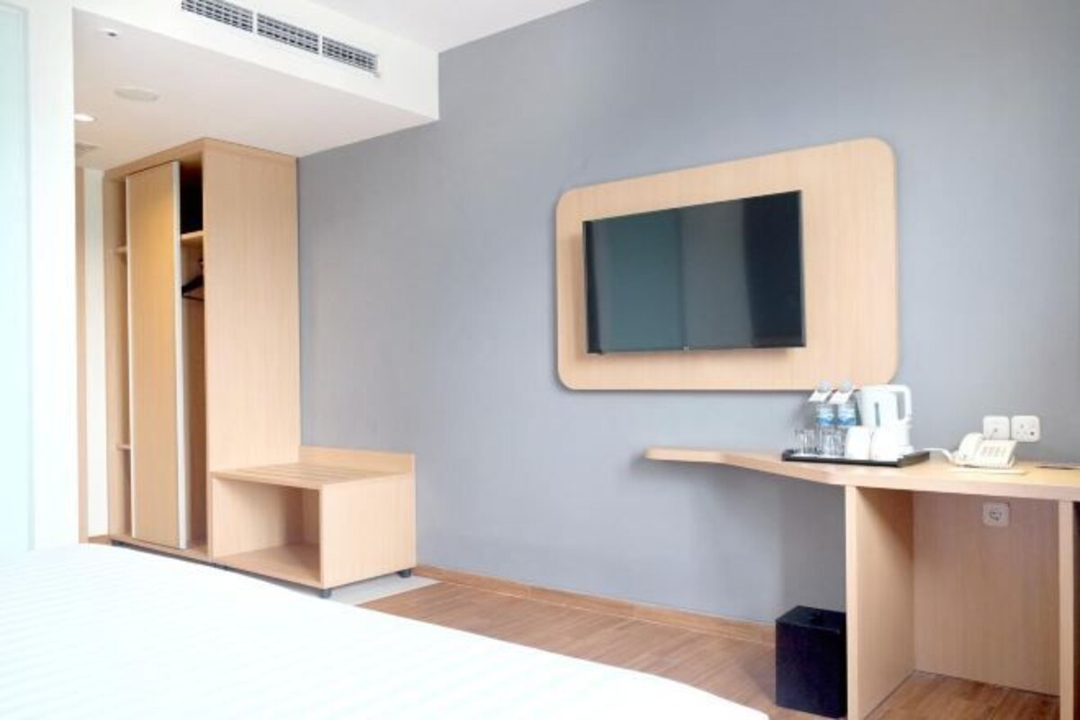 Ett modernt hotellrum med en garderob i trä, en platt-TV som är monterad på väggen, ett litet skrivbord med en telefon och en kaffemaskin samt en papperskorg.