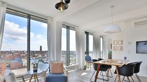 A modern living room with a large window, a dining area with a table and chairs, and a view of the cityscape.