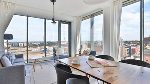 A modern room with a large dining table, chairs, and a sofa. There are large windows offering a view of the city.
