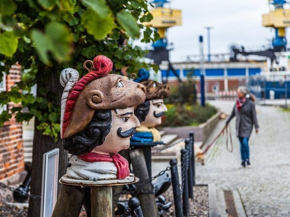Une rangée de statues aux traits du visage et aux détails vestimentaires distinctifs, se dressant sur fond de structures industrielles et d’un chemin.