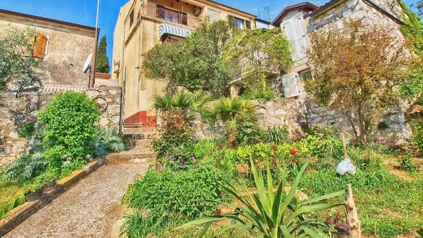 Un sendero estrecho que lleva a un edificio con balcón, rodeado de abundante vegetación y un muro de piedra.