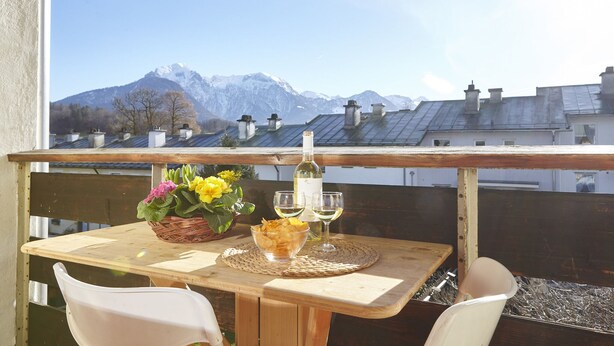 Una mesa de madera con una botella de vino, dos copas, un cuenco de patatas fritas y una cesta de flores en un balcón con vistas a una montaña.