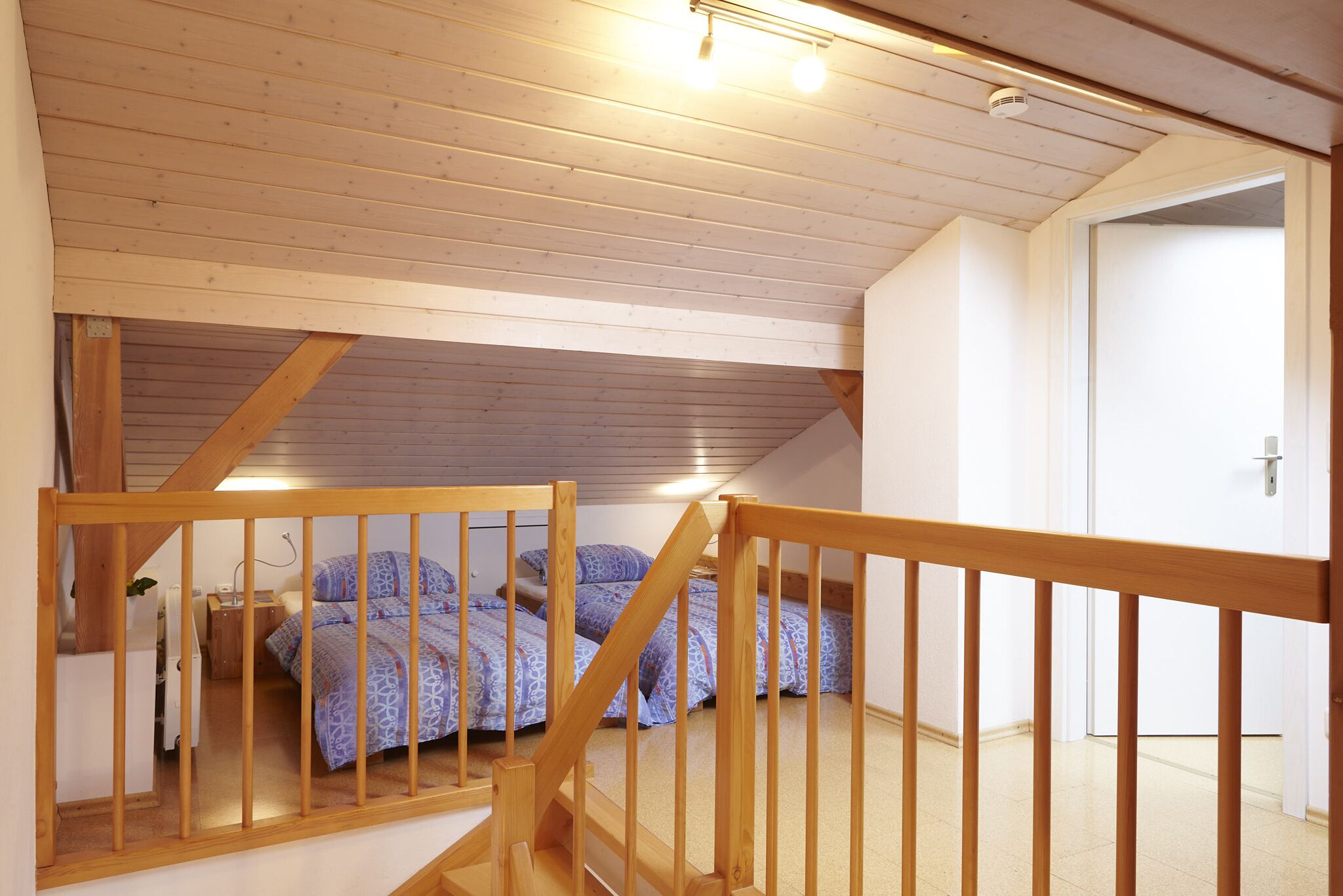 A bedroom with a wooden ceiling, a staircase with a wooden railing, and a bed with patterned bedding.