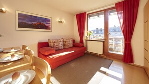 Una sala de estar acogedora con un sofá rojo, una mesa de comedor con platos y cubiertos, un cuadro en la pared y una ventana con cortinas rojas.