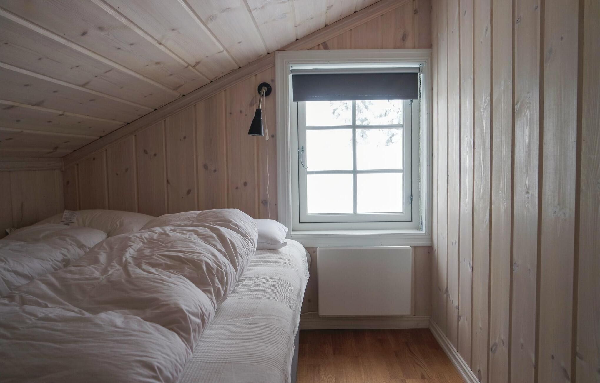 A small, single-bed room with wooden walls and a sloped ceiling. A window with a black blind is on the wall, and a radiator is below it.