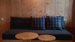 A dark-colored sofa with plaid cushions against a wooden wall, accompanied by two round wooden tables. - Ferienwohnung Nr 1,  2 Pers. 50 qm (Schleching)