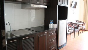 A compact kitchen with dark cabinets, a stainless steel sink, and a small refrigerator. There is a dining area with wicker chairs and a flat-screen TV mounted on the wall. - K&J Apartment (Pattaya)