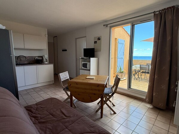 Habitación compacta con kitchenette, comedor con mesa y sillas, televisor y puerta corrediza de vidrio que da a un patio exterior con mesa y sillas.