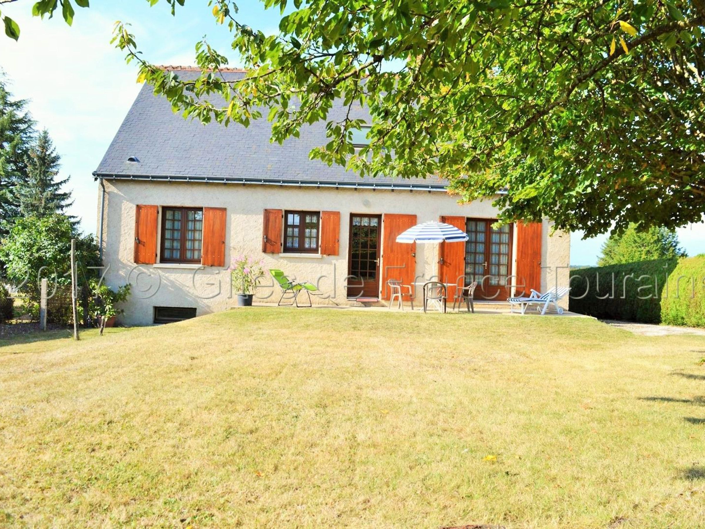Ein zweigeschossiges Haus mit geneigtem Dach, einer Veranda mit einer Bank und einem kleinen Garten mit Rasenfläche und einigen Blumentöpfen.