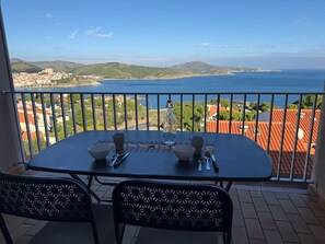 A balcony with a table set for two, overlooking a coastal town and the sea.