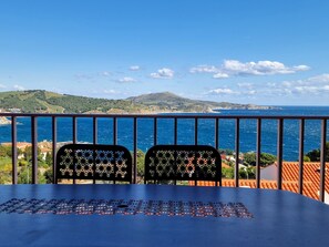 A balcony with two chairs and a table, overlooking a coastal view with hills and the sea.