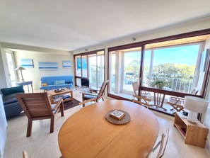 A spacious living area with a large wooden table, comfortable seating, a blue sofa, and a balcony with a view.