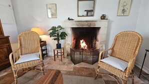 Een gezellige woonkamer met twee rieten fauteuils, een houten salontafel, een open haard en een kleine bijzettafel met een lamp en een plant.