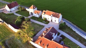 Luchtbeeld van een landgoed met meerdere gebouwen, waaronder een groot huis met een rode dakbedekking en kleinere constructies, omringd door groene velden en een verharde weg.