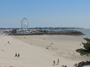 Uma praia com uma roda-gigante ao fundo, um paredão rochoso e algumas pessoas espalhadas pela areia.