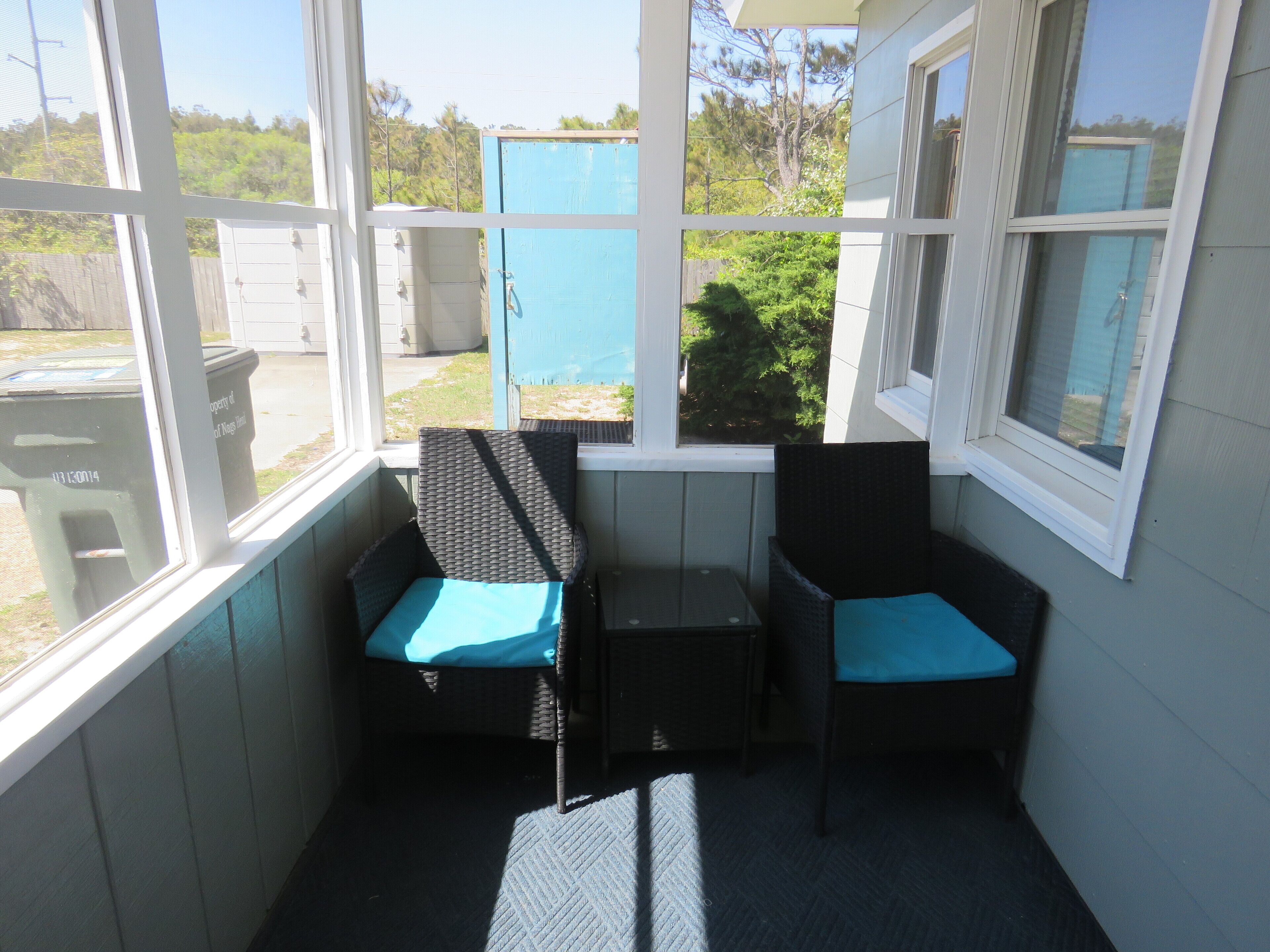 A small enclosed porch with two wicker chairs and a table, large windows, and a view of the outdoors.