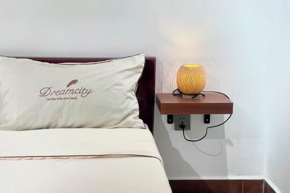 A bed with a pillow that reads "Dreamcity Let me take your hand," and a wall-mounted lamp on a wooden shelf.