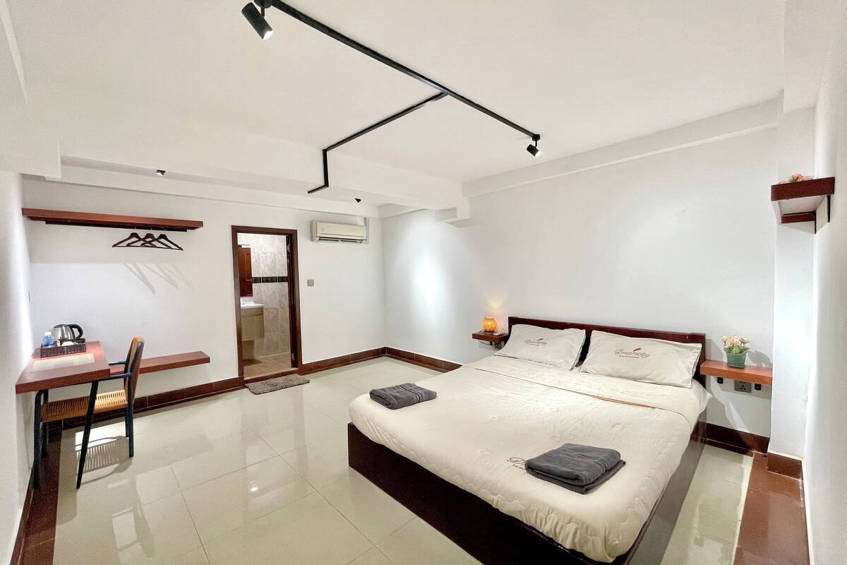 A modern hotel room with a large bed, a desk with a chair, a bedside table with a lamp, and a wardrobe.