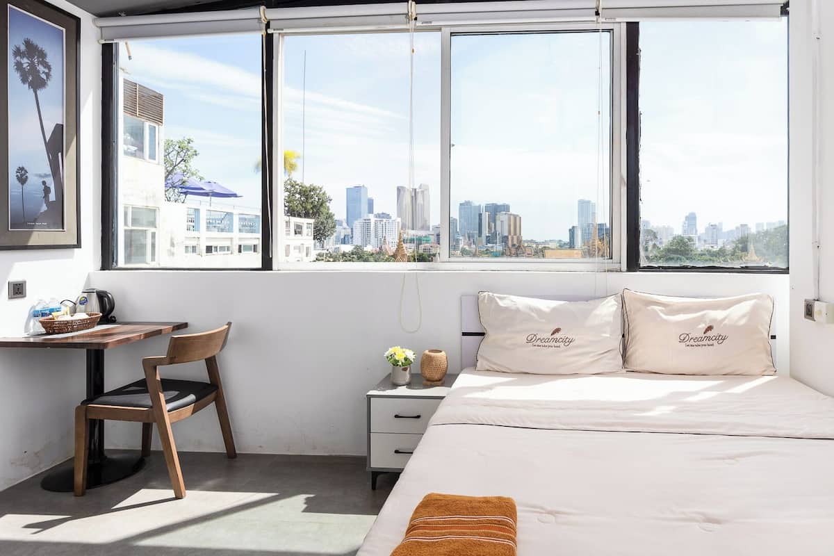 A bedroom with a city view, a desk, a chair, a nightstand, and a bed with pillows.