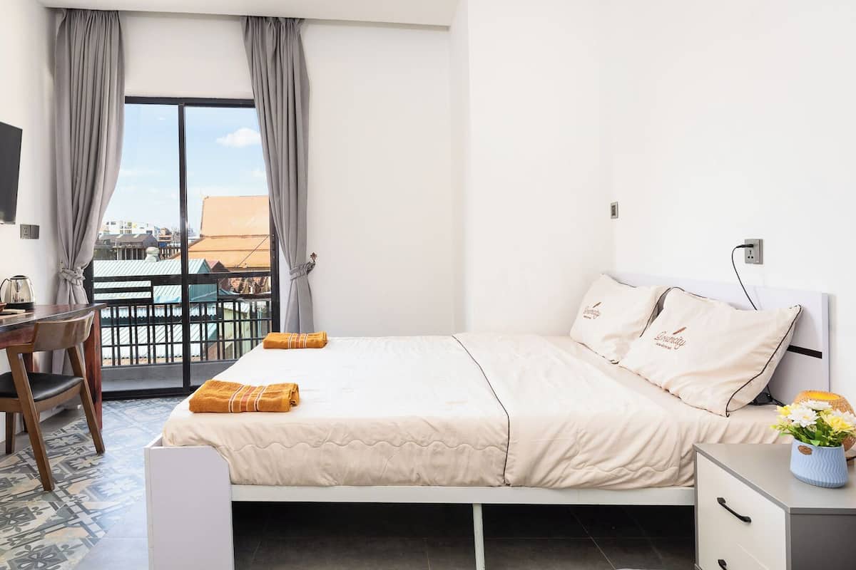 A hotel room with a bed, a desk, a chair, a TV mounted on the wall, and a balcony with curtains.