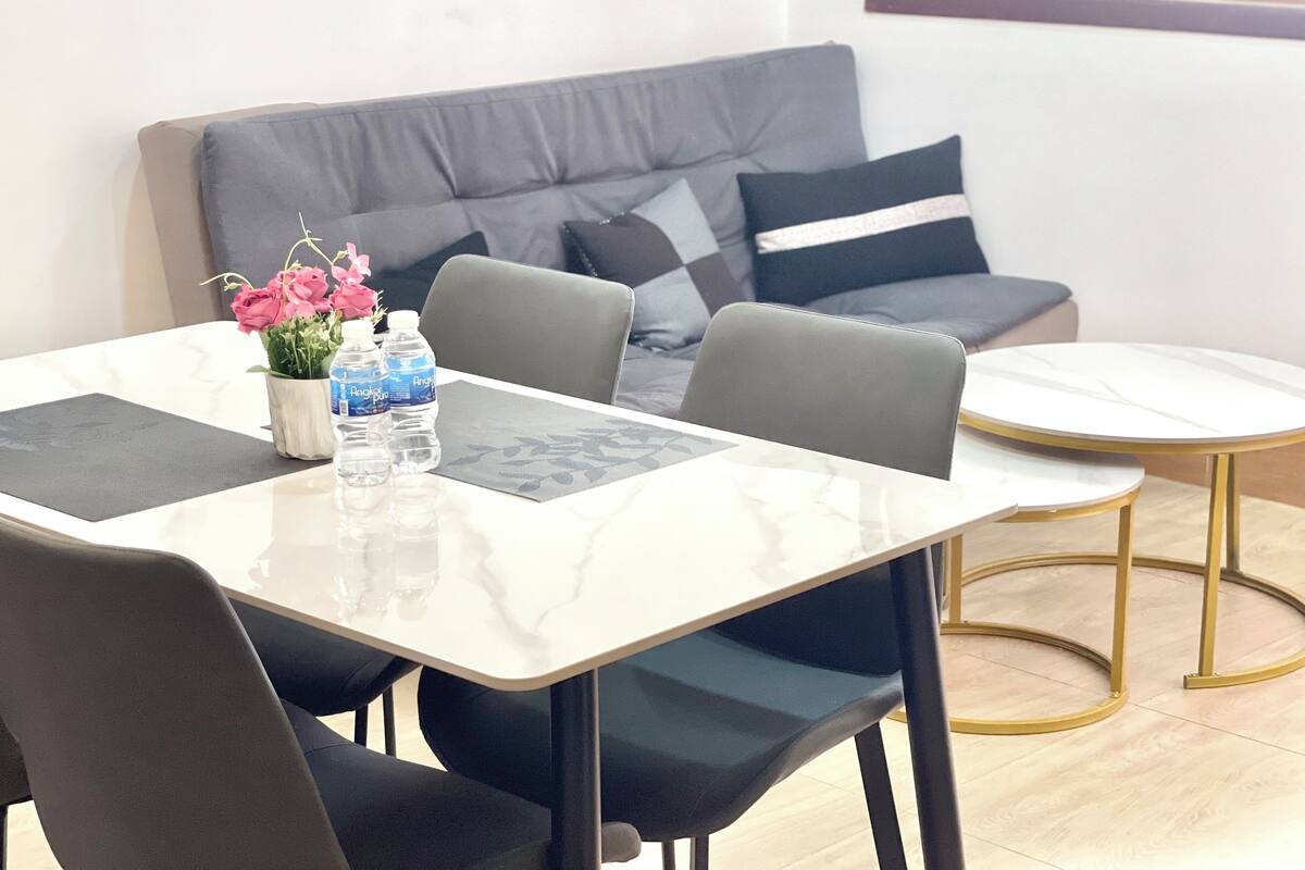 A modern living room with a grey sofa, a small round table, and a rectangular dining table with chairs. There are two bottles of water and a vase with flowers on the dining table.