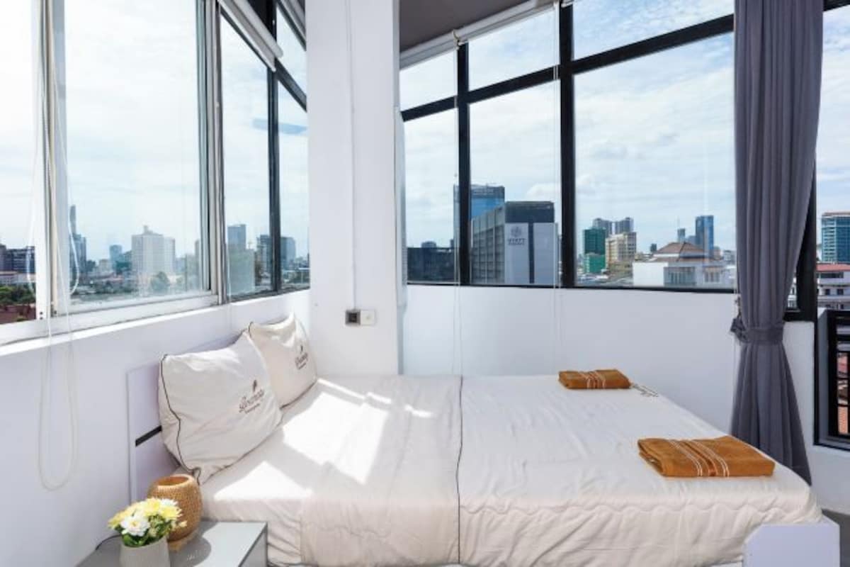 A modern hotel room with a large bed, a bedside table with a vase of flowers, and a view of the cityscape through large windows.
