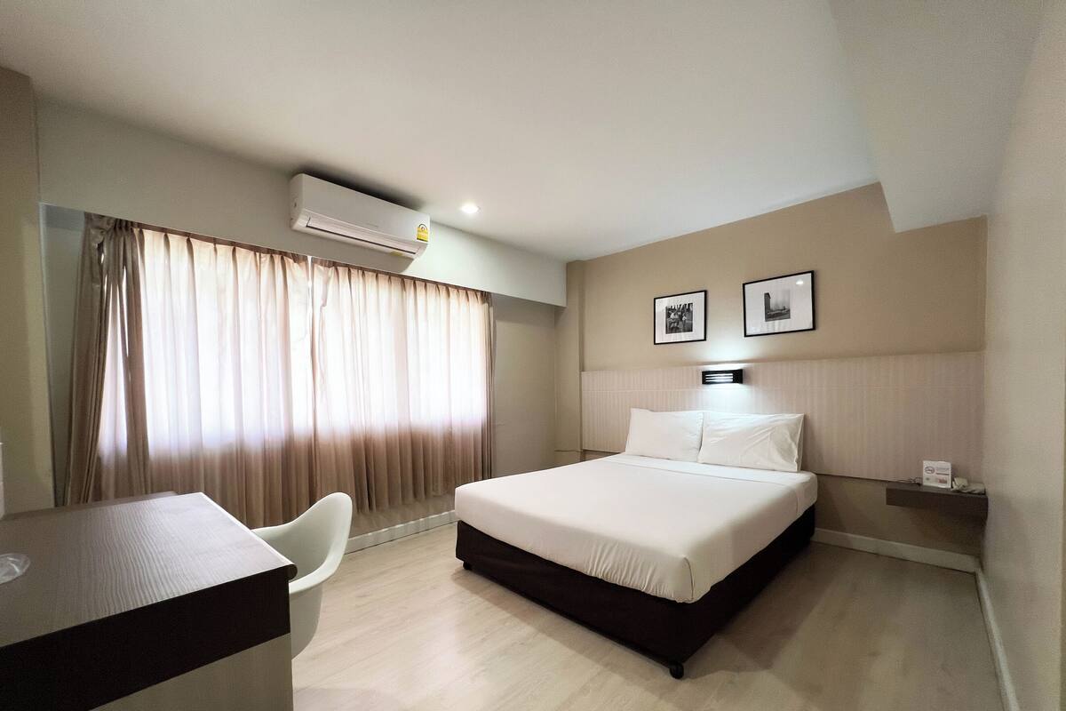 Quarto de hotel com uma cama, uma escrivaninha, uma cadeira, unidade de ar condicionado e duas imagens emolduradas na parede.