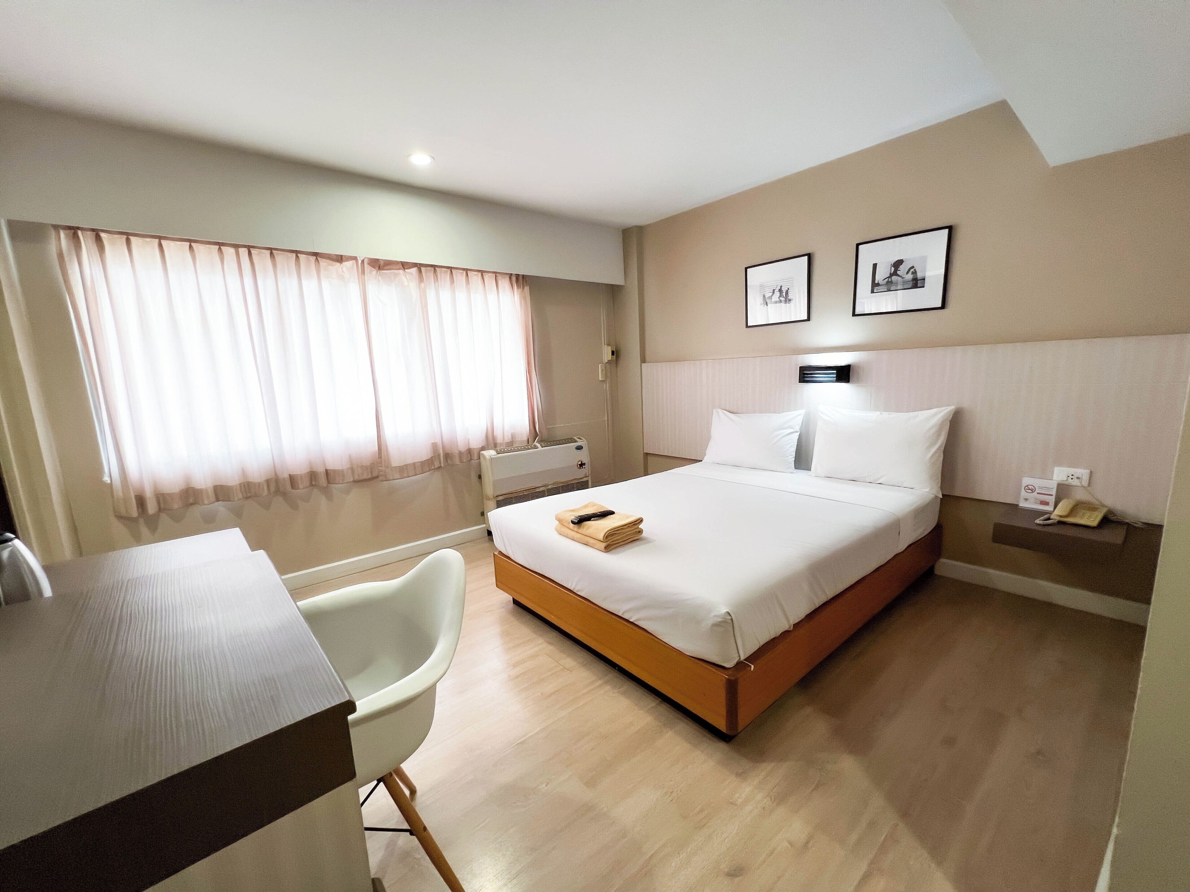A hotel room with a bed, a desk, a chair, a bedside table with a phone, and a window with curtains.