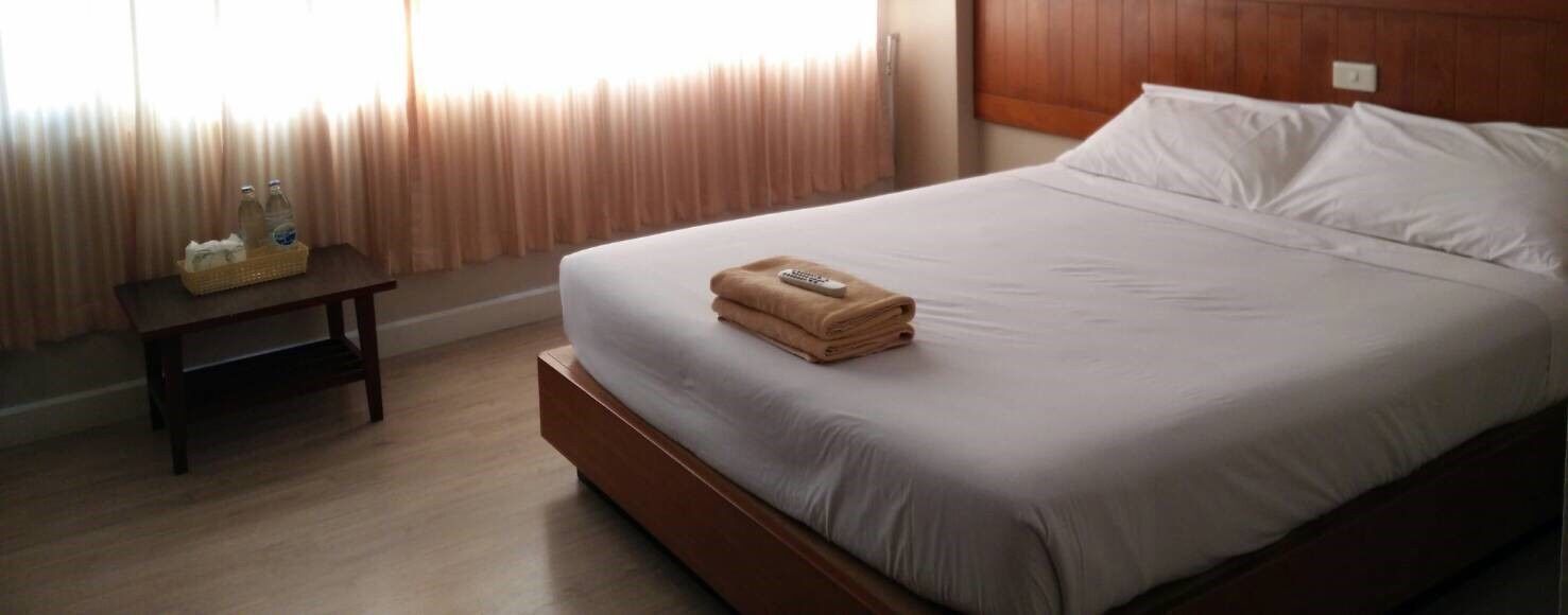 Un lit bien fait, avec une télécommande à portée de main, une petite table sur laquelle se trouve un panier, et une fenêtre avec des rideaux.