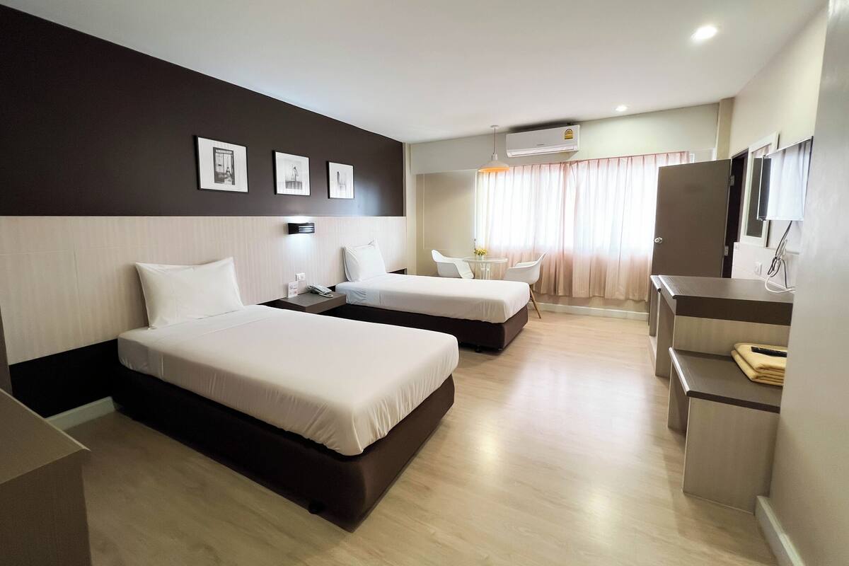 Quarto de hotel com duas camas, uma escrivaninha, uma cadeira e uma mesinha.