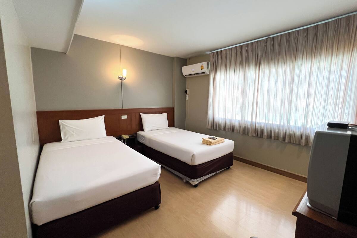 Quarto de hotel com duas camas, uma escrivaninha, uma cadeira, um abajur, um aparelho de ar condicionado e uma janela com cortinas.
