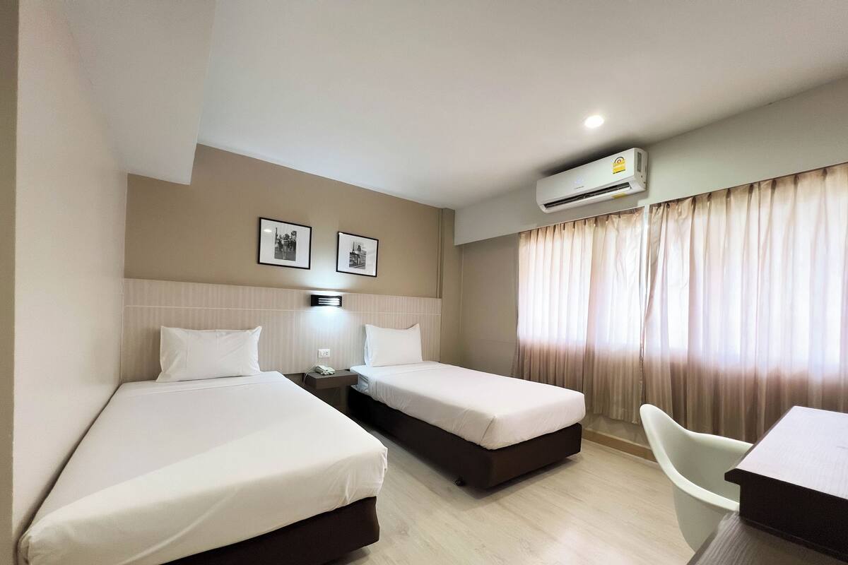 Quarto de hotel com duas camas, uma escrivaninha, uma cadeira, um aparelho de ar condicionado e duas imagens emolduradas na parede.