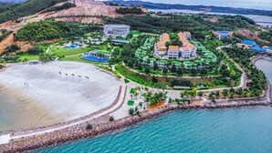 Vista aérea de un complejo turístico con una amplia playa de arena, una piscina y una zona ajardinada con edificios y vegetación.