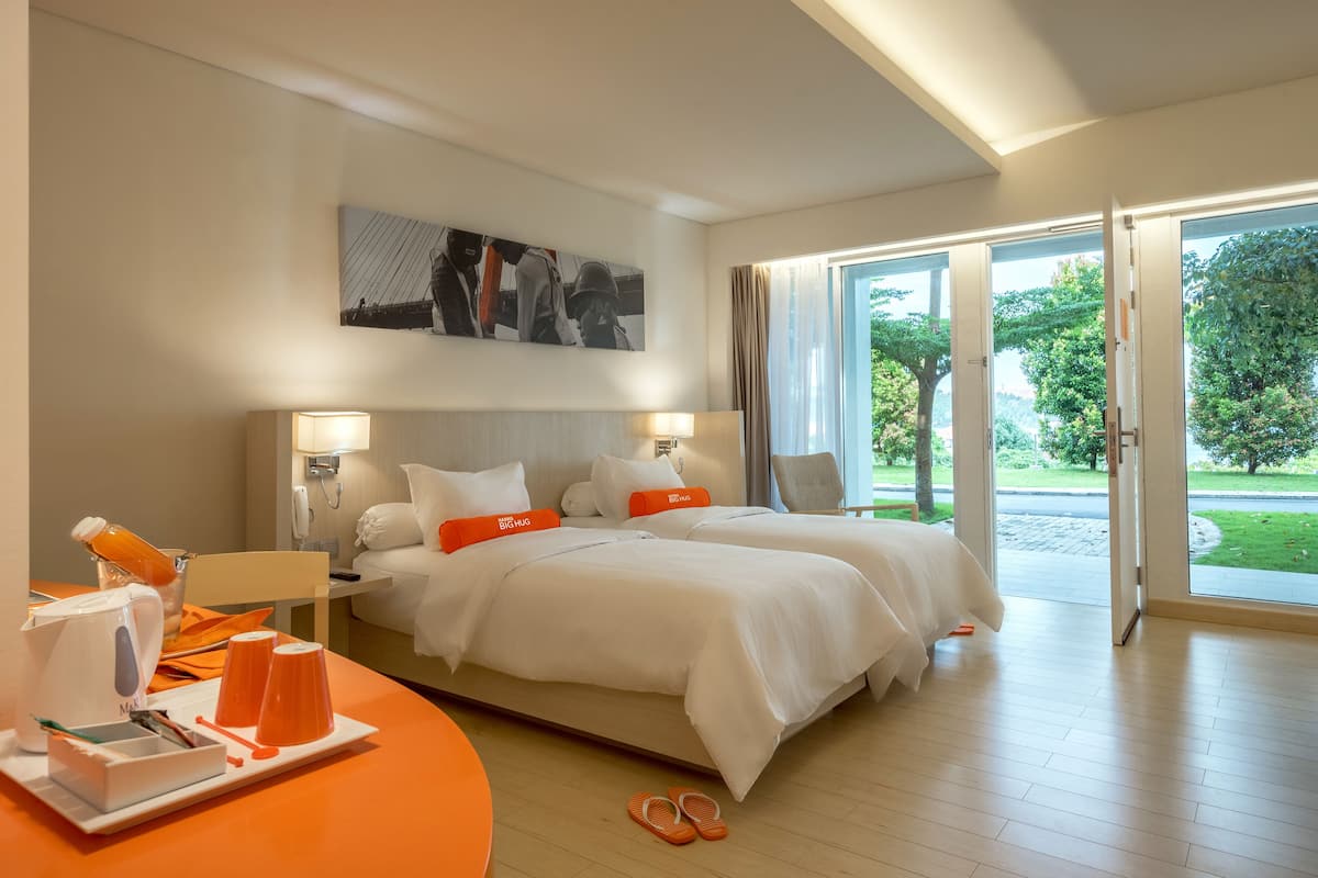 A hotel room with two beds, a large window, and a view of greenery outside.