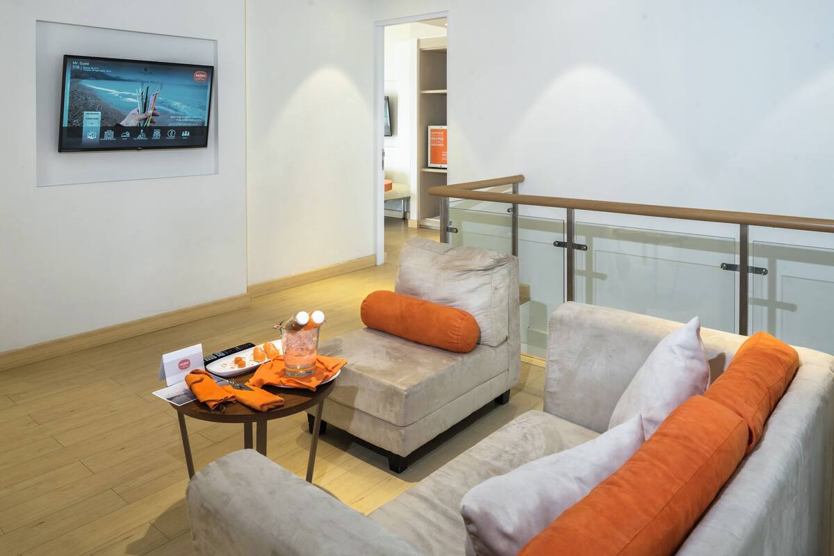 A modern living room with a sofa, armchair, and a small table with a glass. A TV is mounted on the wall, and a balcony railing is visible.
