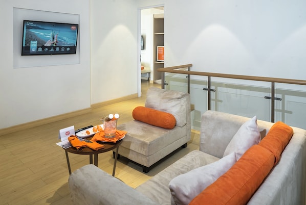 Un salon moderne avec un canapé, un fauteuil et une petite table sur laquelle se trouve un verre. Un téléviseur est fixé au mur, et on aperçoit la rambarde d’un balcon.