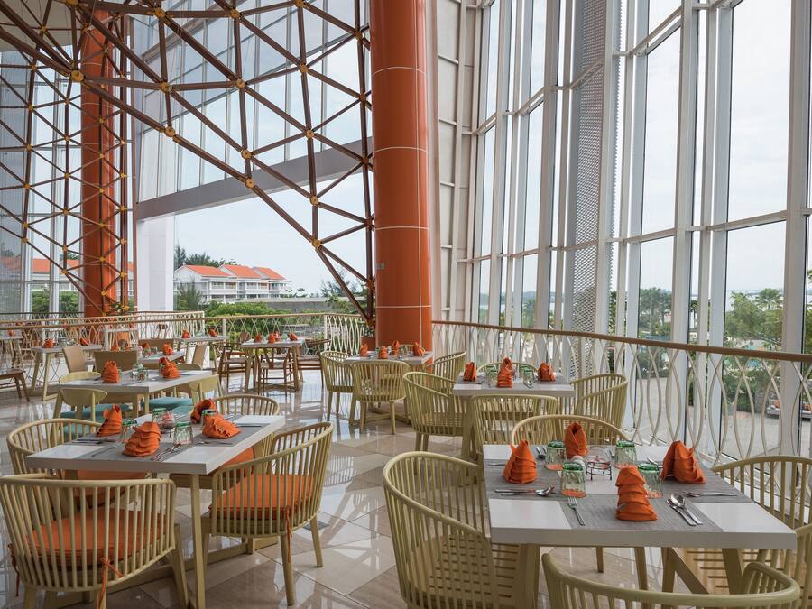 Un amplio espacio al aire libre para comer, con varias mesas y sillas, ventanas grandes y vistas a edificios al fondo.