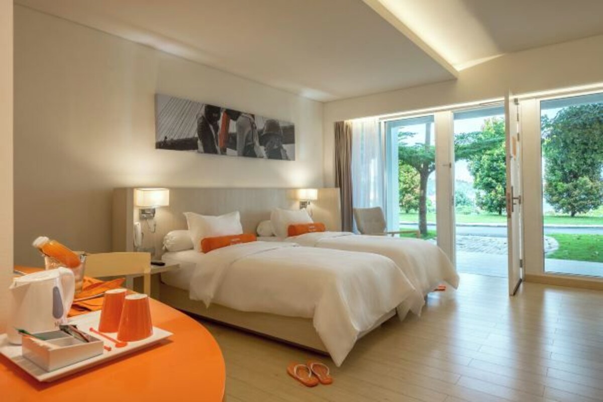 A hotel room with two beds, a large window with a view of greenery, and a dining area with orange chairs and a table.