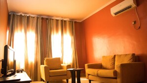 A room with two yellow armchairs, a small round table, a television, and an air conditioning unit on the wall. - The Stonehouse Hotel (Lekki)