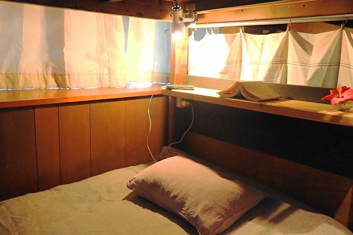 Una cabaña con una cama, una ventana con cortinas, una estantería con libros y una flor, y una pared de madera.