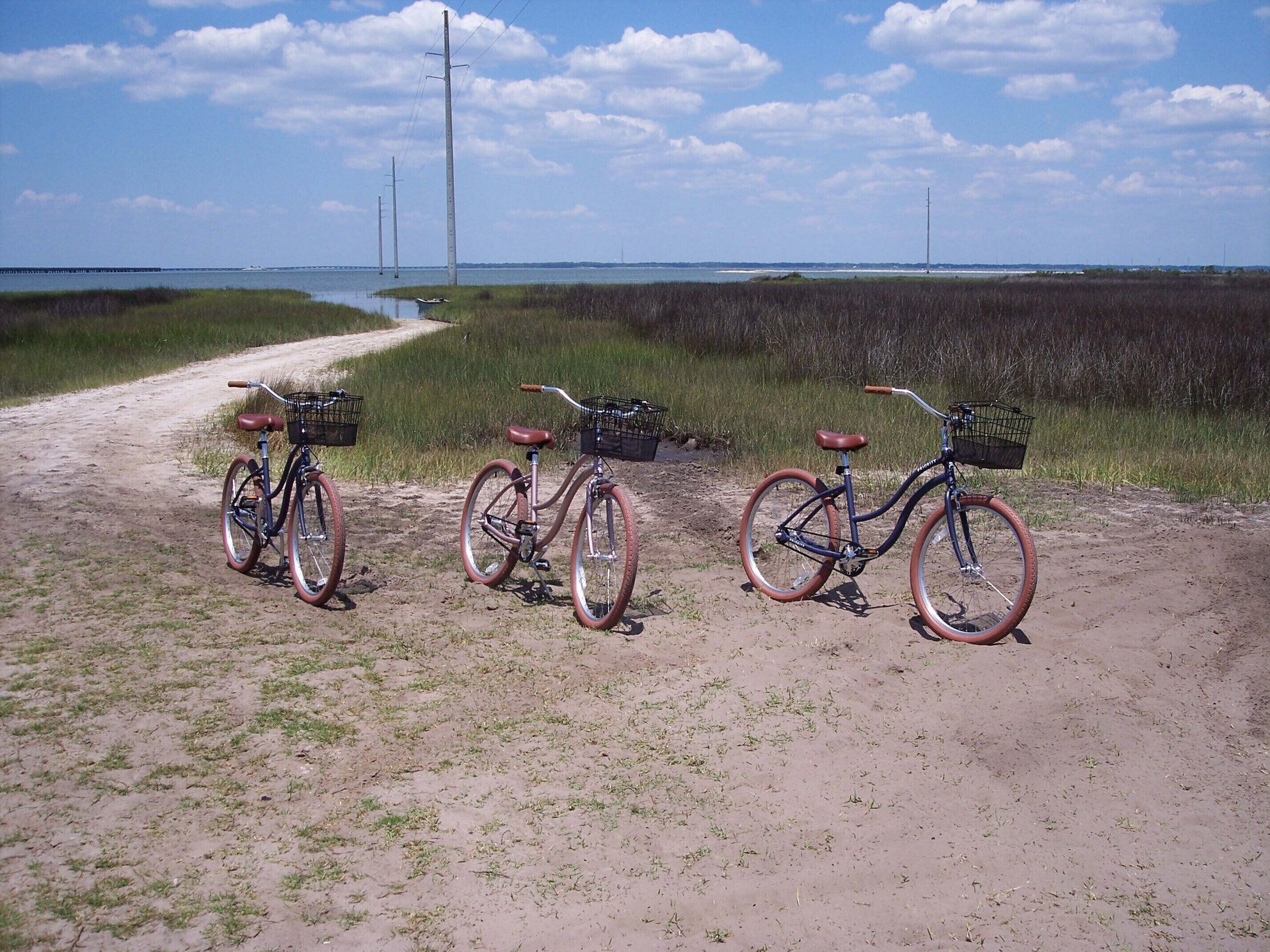 Three bicycles parked on a dirt path with a grassy field and a body of water in the background.