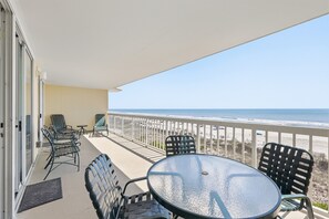 A balcony with a glass table and chairs, overlooking a beach.